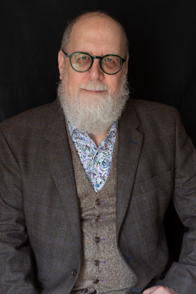 An image of a balding man with white beard and glasses, wearing a shirt, jacket and waistcoat (vest)